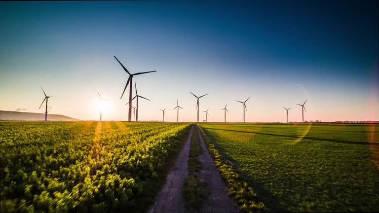 Flight over a field road surrounded by wind turbines back lit by warm sunlight at sunset. Great for alternative and renewable energy themes. Shot in North Rhine Westphalia in Germany.