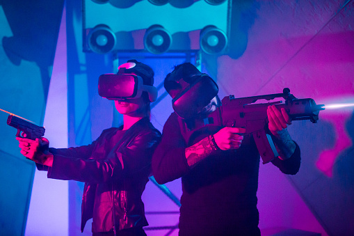 The young woman and young man are playing a virtual reality shooting game. They are using headset (VR glasses on heads) and handguns. The woman and man are aimining from handguns. They standing back to back. Studio shooting with multi colored (red and blue) lighting