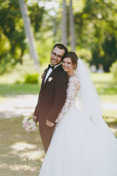 beau mariage photosession. le marié en costume noir et son épouse dans une robe en dentelle blanche avec un long panache, le voile et sourire de bouquet et embrasser dans un grand jardin verdoyant sur une journée ensoleillée weathery - veil bride lace married photos et images de collection