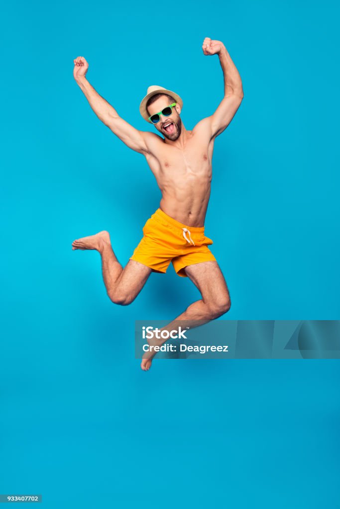 Fullbody, full length portrait of attractive, successful, cheerful, foolish, comic ladies' man in yellow shorts, jumping with open mouth and raised hands, isolated on blue background Men Stock Photo