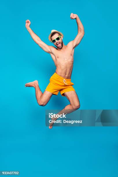 Fullbody Full Length Portrait Of Attractive Successful Cheerful Foolish Comic Ladies Man In Yellow Shorts Jumping With Open Mouth And Raised Hands Isolated On Blue Background Stock Photo - Download Image Now