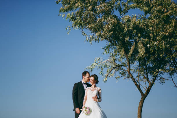 beau mariage photosession. beau marié dans un costume noir et une jeune mariée en robe de dentelle blanche avec coiffure exquise promenade autour du terrain contre le ciel et le grand arbre - veil bride lace married photos et images de collection