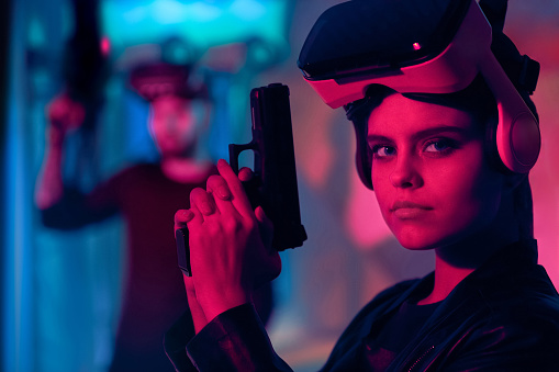 The young woman in the virtual reality headset (VR glasses) on her head. She is holding the game handgun in her hands. The woman looking at the camera. The young man with virtual reality game equipment stands behind the woman. Studio shooting with multi colored (red and blue) lighting