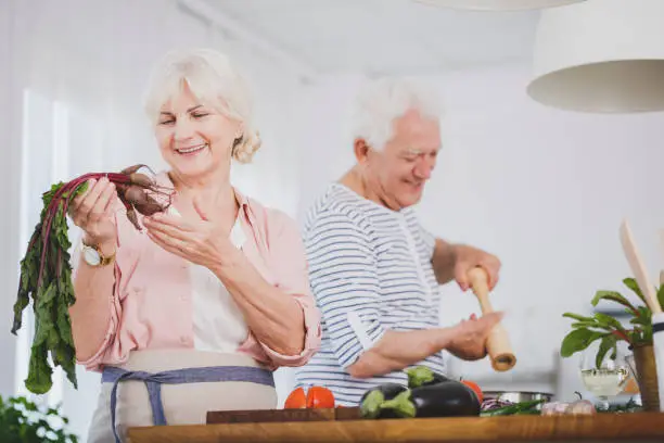Senior woman holding small beetroot and an older man seasoning food, preparing a healthy vegan meal together