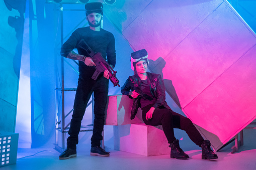 The young woman and young man are using a virtual reality equipment. They are posing together (the woman is sitting and the man standing next to her). The couple is looking at the camera. Studio shooting with multi colored (red and blue) lighting