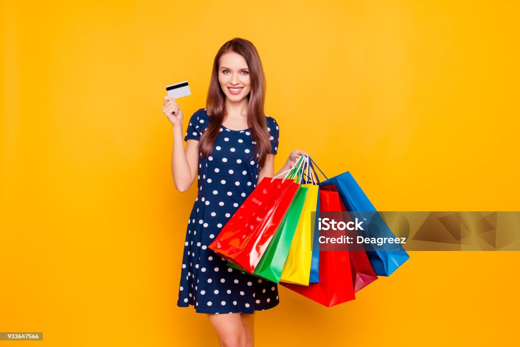 Charming pretty attractive sexy girl showing credit card and holding many colorful packages in hand, it is comfortable to use bank card for shopping, standing over yellow background Shopping Bag Stock Photo