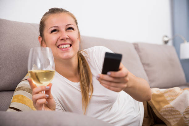 istock Young woman is sitting with white wine and watching TV 933661886