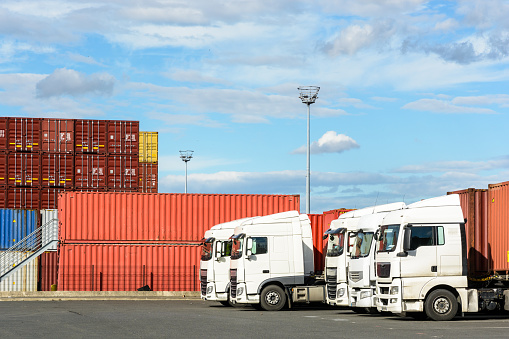 A row of white semi-trailer trucks parked on the container storage platform of the intermodal terminal of a river port with containers in the background.