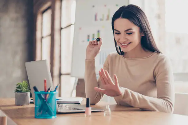 Photo of Portrait of fashionable stylish modern dreamy careless excited satisfied elegant attractive charming gorgeous financier applying varnish on nails sitting at the desktop reception room