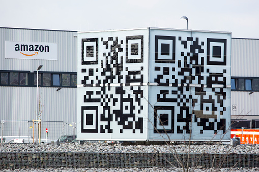 Facade and logo of new logistics center of amazon in Raunheim-Moenchhof, Germany. In the foreground a large qr-code cube made out of plastic. Amazon (Amazon.com, Inc.) is an American electronic commerce and cloud computing company and the largest Internet retailer in the world as measured by revenue and market capitalization.