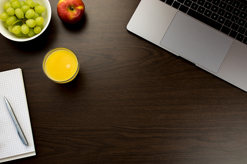 Desk in the office with a notebook next to grapes, an apple and a glass of orange juice, notepad with pen, space in the middle - wooden table