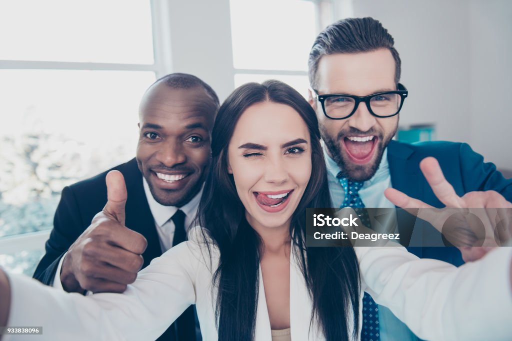 Three comic, funny, crazy partners, colleagues having fun, men with bristle showing like symbol and two fingers, peace sign with hands, girl shooting selfie and gesture tongue out, wink with one eye Working Stock Photo