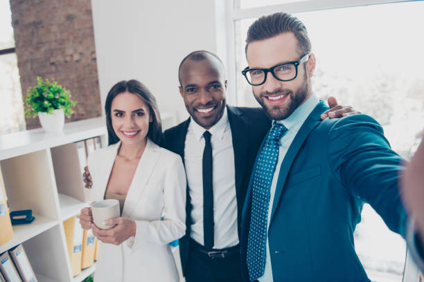 istock Self portrait of three partners, bearded man in glasses making selfie on front camera with his colleagues in classic suits, holding mug with tea in hand, standing in work place, station 933843740