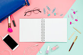 Frame flatlay with woman's blue bag, glasses, smartphone with black copyspace, cosmetics, stationary supples and notebook with blank white page. Pastel background, business mockup, women bag contents