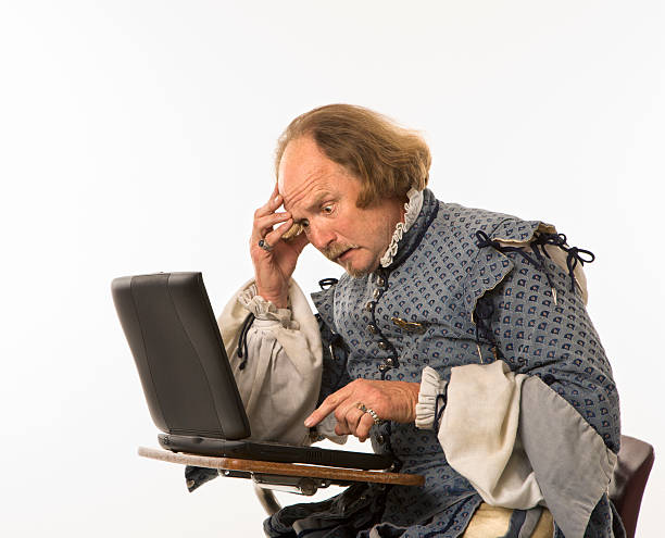 Shakespeare using laptop. William Shakespeare in period clothing sitting in school desk with laptop computer and hand to head looking perplexed. william shakespeare photos stock pictures, royalty-free photos & images
