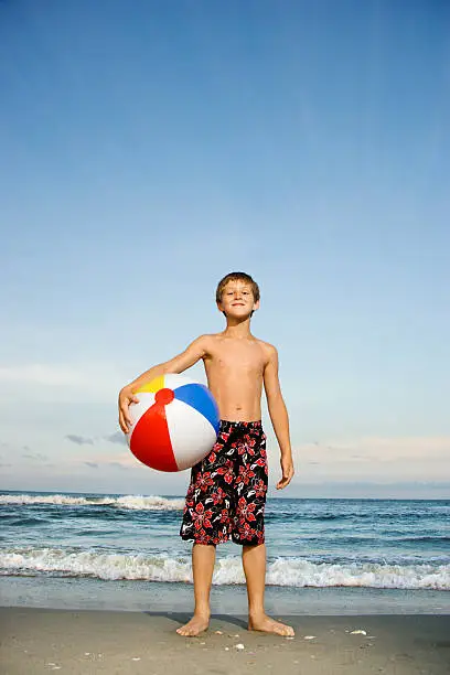 Caucasian pre-teen boy holding beachball on beach.