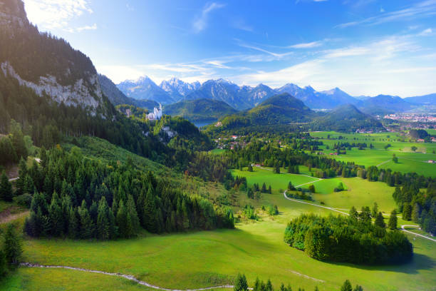 famous neuschwanstein castle visible in the distance, located on a rugged hill above the village of hohenschwangau in southwest bavaria, germany - international landmark sunny lake sky imagens e fotografias de stock