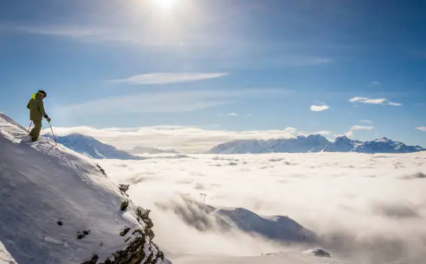 A skier in the Swiss ski resort of Verbier standing on top of a cliff above a sea of clouds, looking towards Mont Blanc and Chamonix.