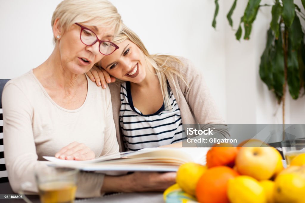 Young woman resting her head on mother's shoulder while looking at family photo album Happy senior mother and her daughter looking at family photo album while sitting at a dining table. Daughter's head resting on mother shoulder. Happy family moments at home. Daughter Stock Photo