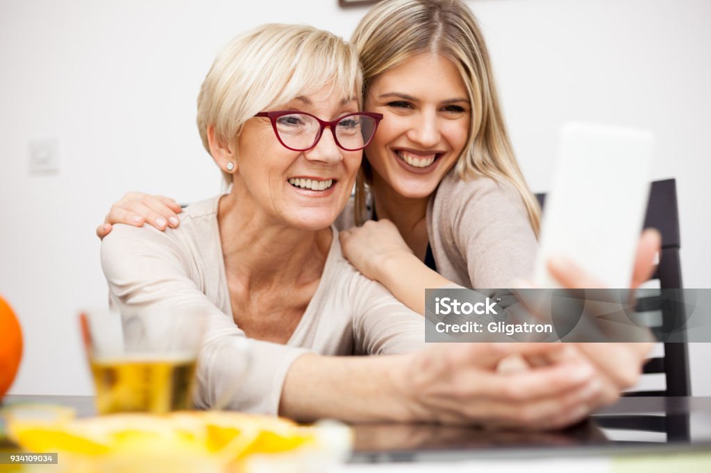 Senior woman and her daughter having fun and posing for a selfie Senior mother and her daughter smiling and posing for a selfie while sitting by dinner table in bright room. Mother holding phone. Happy family moments at home. Women Stock Photo