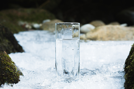 Transparent glass glass with drinking mountain water in winter stands on an icy crust against the background of a clean, frosty river bank. The concept of drinking mountain drinking mineral water and the production of mineral ecologically clean drinking water. The concept of using environmentally friendly products.