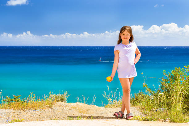Cute happy girl in a summer dress with a ripe yellow lemon in hand posing against background of ultramarine turquoise Cretan sea. Cliff above Livadi beach in resort village Bali, Crete, Greece
