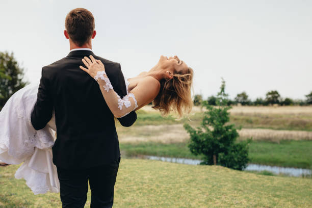 Full length shot of a young mixed race couple standing together on grass celebrating their wedding day. Bride and groom standing face to face and touching foreheads while posing for wedding photo shoot