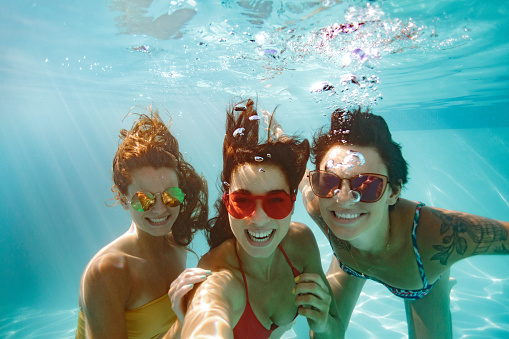 Cheerful women friends swimming underwater in pool taking selfie. Underwater selfie of happy females in pool.