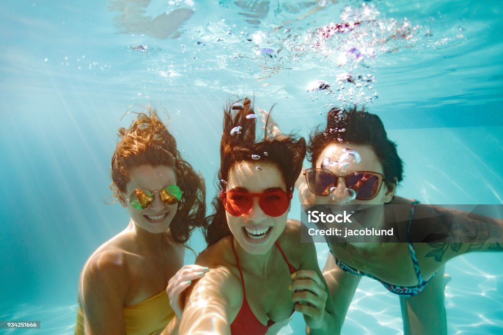 Cheerful friends making selfie underwater in pool Cheerful women friends swimming underwater in pool taking selfie. Underwater selfie of happy females in pool. Swimming Pool Stock Photo