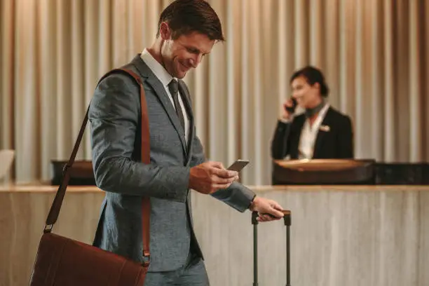 Businessman walking in hotel lobby with suitcase and using his smart phone. Male business traveler in hotel hallway with cellphone and luggage.
