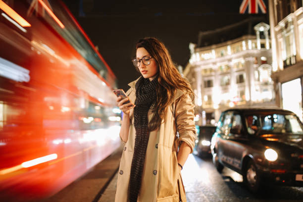 Portrait of a young woman on the busy streets of London downtown in the evening, texting for a cab Vintage toned portrait of a young woman, wearing a trench coat, walking on the busy streets of London in the evening. Going home after work, texting on her cellphone, trying to find Uber or a cab. london fashion stock pictures, royalty-free photos & images