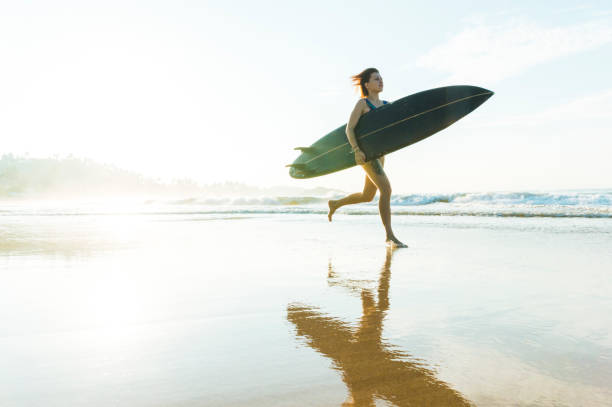 istock Young woman runs along beach with surfboard 934280630