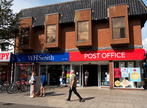 On a sunny summer day, people and a dog outside a branch of the newsagent chain WH Smith in Lowestoft, Suffolk, Eastern England. In recent years, many British post offices have closed and WH Smith has taken on the franchise, as is the case here.