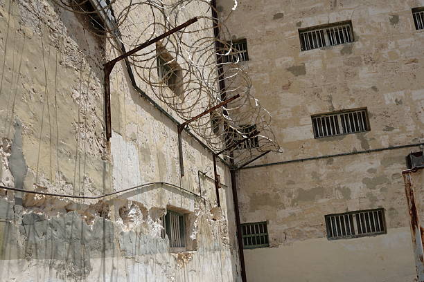 Row of small prison cells with bars in front. Inside the cell is a bed and toilet.