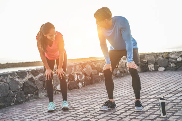 fit couple taking a rest after fast running workout - joggers training outdoor at sunset - focus on man face - fitness, sport, wellness, workout, gym and healthy lifestyle concept - stretch beach imagens e fotografias de stock
