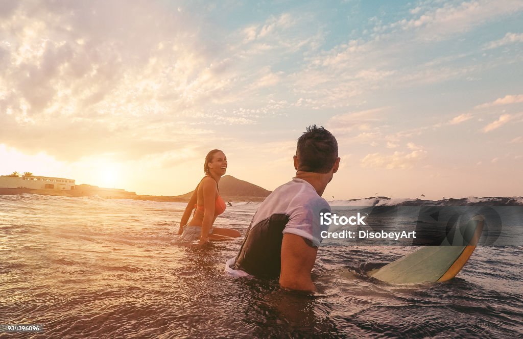 Fit couple surfing at sunset - Surfers friends having fun inside ocean - Extreme sport, travel, healthy lifestyle and vacation concept - Focus on man head Surfing Stock Photo