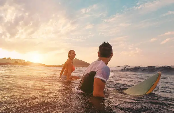 Photo of Fit couple surfing at sunset - Surfers friends having fun inside ocean - Extreme sport, travel, healthy lifestyle and vacation concept - Focus on man head