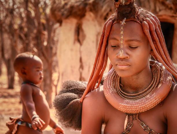 vista ravvicinata che mostra i gioielli tradizionali, l'acconciatura e la pasta di pelle ocra di una donna tribale himba, con un bambino in sottofondo. - necklace horizontal waist up outdoors foto e immagini stock