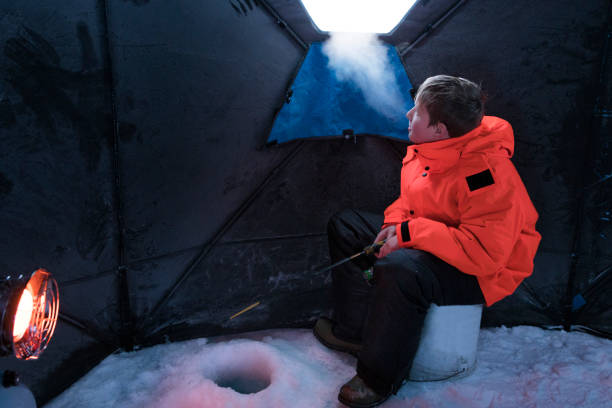A 13 year old boy sitting on a bucket in a canvas hut can see his frosty breath while ice fishing on a subzero winter day in northern Minnesota, USA