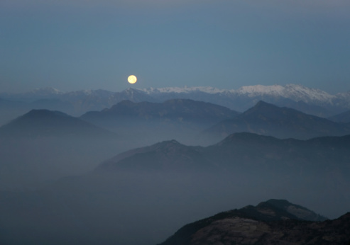 A breathtaking image of a full moon set amongst a mountainous and tree-filled landscape