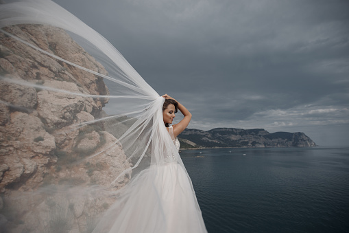 Beautiful bride stands on a cliff above the sea in a glamorous white wedding dress view of veil.Romantic beautiful bride in white dress posing on the background sea. Happy bride in luxury dress posing on the beach.Happy Wedding Day.