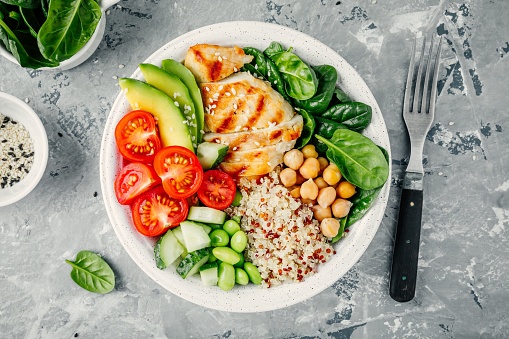 Buddha bowl with spinach salad, quinoa, roasted chickpeas, grilled chicken, avocado, tomatoes, cucumbers, sesame seeds. Top view