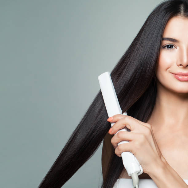 beautiful woman with long straight hair using hair straightener. cute smiling girl straightening healthy brown hair with flat iron on gray background - root hair imagens e fotografias de stock