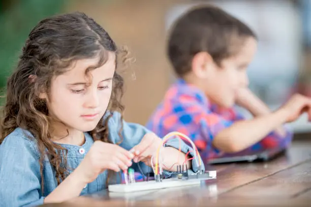 A young brother and sister are at school, working on a science project.The girl is working with wires and electricity in the foreground.