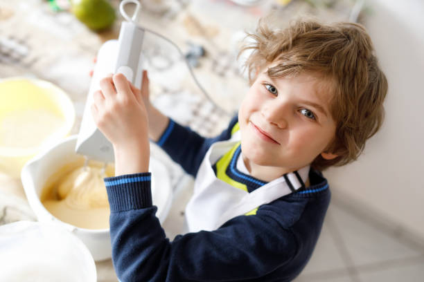 beautiful funny blond little kid boy baking chocolate cake and tasting dough in domestic kitchen - cake making mixing eggs imagens e fotografias de stock