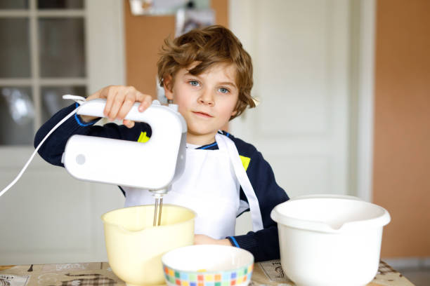 beautiful funny blond little kid boy baking chocolate cake and tasting dough in domestic kitchen - cake making mixing eggs imagens e fotografias de stock