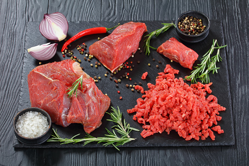 fresh beef forcemeat and pieces of beef filet on black slate plate on black wooden table with salt, peppercorns, chilli and fresh rosemary, view from above