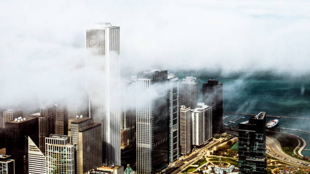 Tall skyscrapers partially hidden by thick fog loom over a modern cityscape. The light reflecting off the buildings adds a mysterious glow to the misty surroundings, enhancing the ambiance.