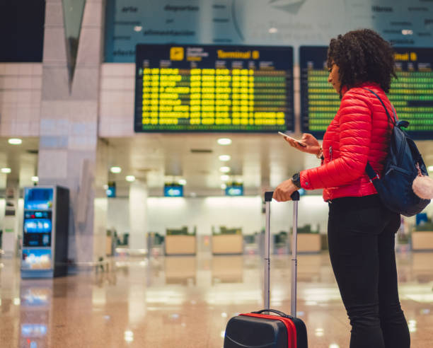 Young mixed race tourist woman checking for flight at airport Young woman at the airport terminal using smartphone heathrow airport stock pictures, royalty-free photos & images