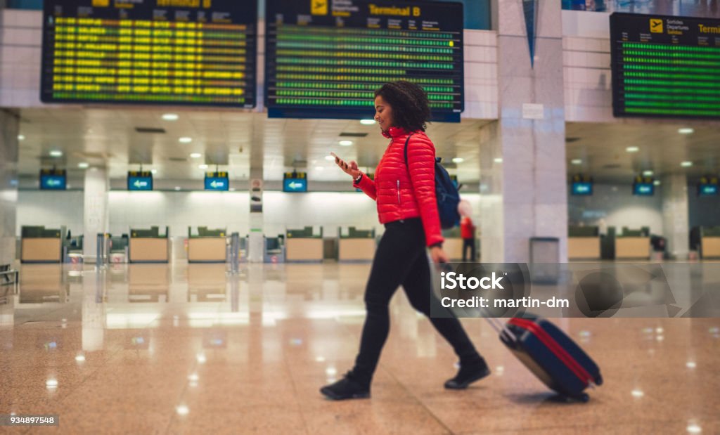 Mixed race woman texting at the airport terminal Young woman with wheeled luggage walking at the airport and texting Airport Stock Photo